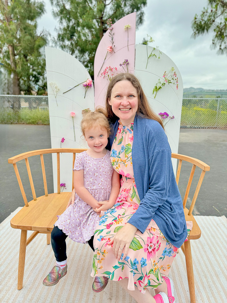Shannon and Beatrix, age 5, in floral dresses, sitting on a bench in front of a backdrop decorated with flowers.