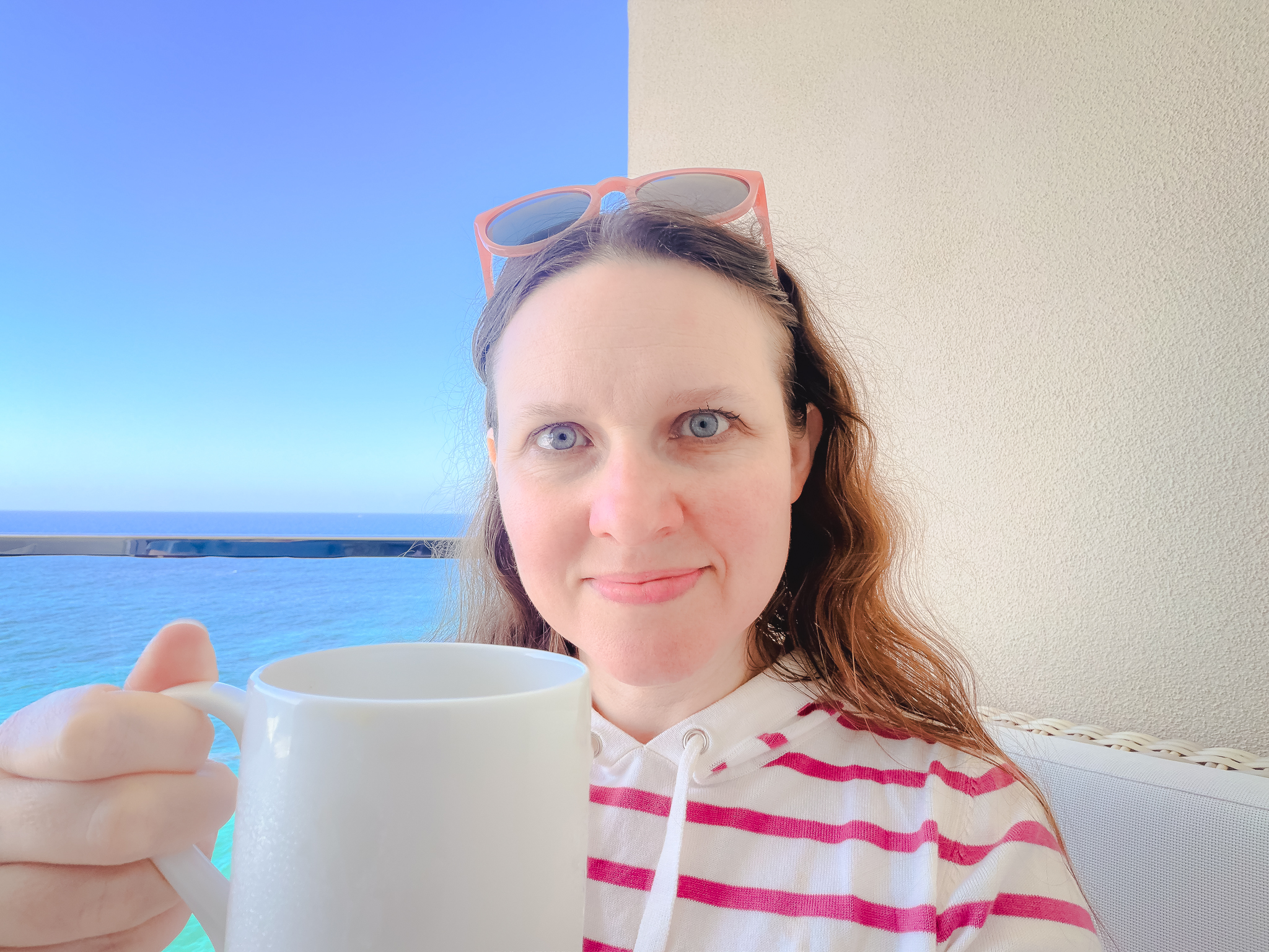 Shannon holding up a coffee cup with the ocean in the background. Shannon is a woman with fair skin, blue eyes, and light brown hair. October 4, 2025